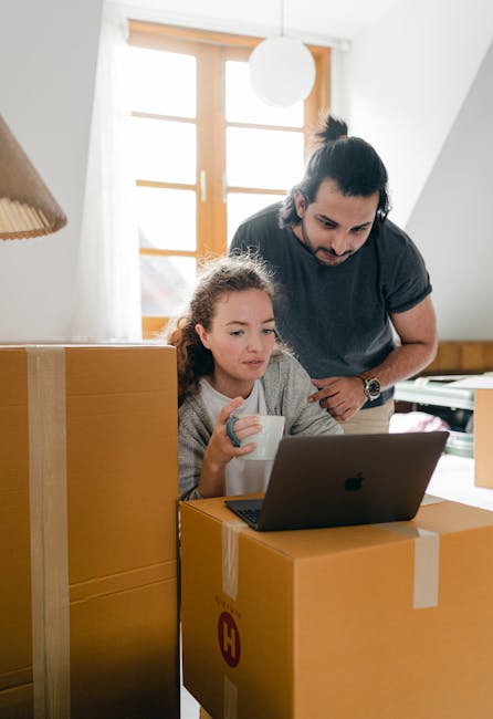 A young girl with curly hair sitting at a desk inside a bright room, drinking from a white mug while looking at a laptop screen placed on top of a large cardboard box. A man, possibly her father, with dark hair tied in a bun, wearing a dark grey T-shirt, is standing beside her, leaning over and observing the laptop. The room features wooden window frames allowing natural light to illuminate the space, and additional cardboard boxes are stacked around, suggesting the process of home packing and packing materials such as cardboard boxes are visible. This scene depicts a home relocation preparation, with furniture and packaging indicating a move in progress, aligned with professional removals and transportation services offered by Haringey Movers.