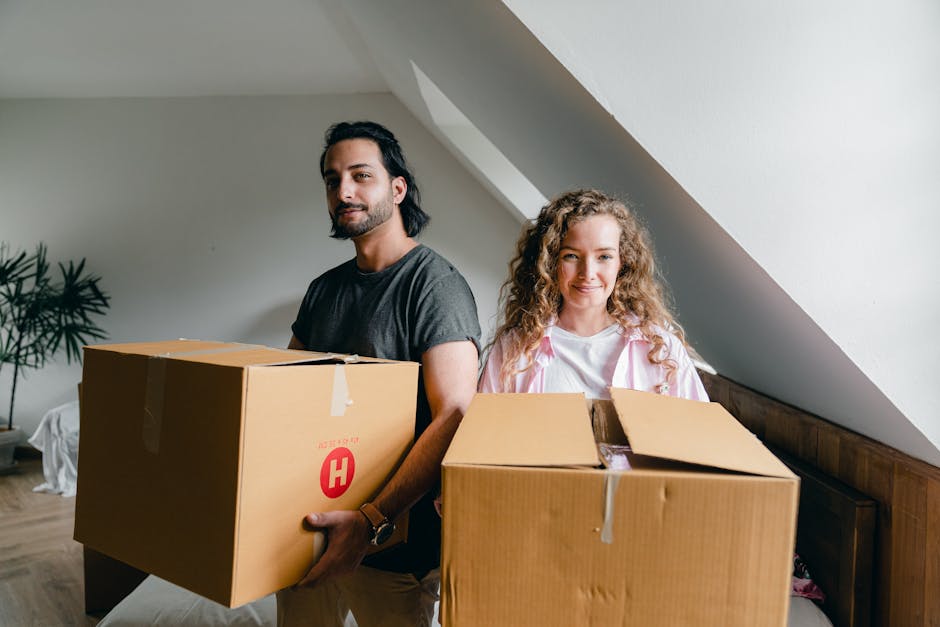 A man and a woman indoors during a home relocation process, each holding a cardboard box filled with packed items. The man, with dark hair and a beard, is wearing a grey t-shirt and is standing on the left side, holding a medium-sized box with a red 'H' logo on it. The woman, with long curly blonde hair, is wearing a white shirt with pink accents and is standing to the right, holding a slightly smaller cardboard box with some packing material visible inside. They are positioned in an attic or loft space, with a sloped ceiling and neutral-colored walls. Behind them, a wooden headboard and a white bedspread are partially visible, along with a potted plant on the left side of the image. The environment is well-lit, suggesting natural light coming through a nearby window, and the scene depicts the loading or packing stage of house removals, with various cardboard boxes prepared for transport by professional movers like Haringey Movers.