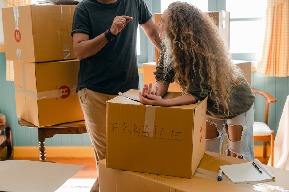 A young girl with curly hair sitting at a desk inside a bright room, drinking from a white mug while looking at a laptop screen placed on top of a large cardboard box. A man, possibly her father, with dark hair tied in a bun, wearing a dark grey T-shirt, is standing beside her, leaning over and observing the laptop. The room features wooden window frames allowing natural light to illuminate the space, and additional cardboard boxes are stacked around, suggesting the process of home packing and packing materials such as cardboard boxes are visible. This scene depicts a home relocation preparation, with furniture and packaging indicating a move in progress, aligned with professional removals and transportation services offered by Haringey Movers.
