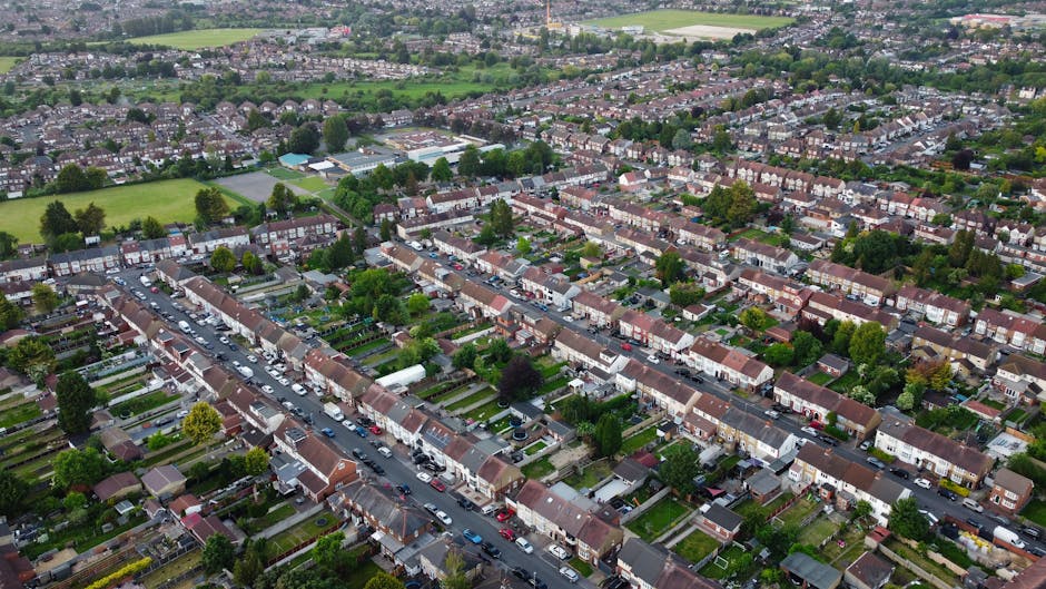 An aerial photograph of a residential neighbourhood in Haringey, showing rows of terraced houses with brick facades and tiled roofs. The street is lined with parked cars on both sides, and there are visible driveways, small gardens, and trees interspersed among the houses. The area includes green spaces and sports fields in the background, with the overall scene captured during daylight on a clear day. This view highlights typical suburban housing layout and the accessibility challenges faced during home relocation and furniture transport, as managed by Haringey Movers' removals services.