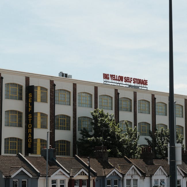 A large multi-storey commercial building with a white exterior and numerous arched windows, some framed with yellow accents, is visible under a partly cloudy sky. Prominently displayed on the rooftop is a red sign reading 'BIG YELLOW SELF STORAGE', indicating a storage facility. Attached vertically to the side of the building is a yellow and black sign that reads 'SELF STORAGE'. In the foreground, there are small residential-style houses with tiled roofs, and a street lamp pole is partially visible on the right side. Trees with green foliage partially obscure the lower part of the building and the houses, and a few staff members or movers are seen carrying or arranging items near the storefronts, suggesting activities related to home or business relocation. The scene captures a typical urban environment where furniture transport and packing activities might be occurring as part of a house removal or moving service provided by Haringey Movers.