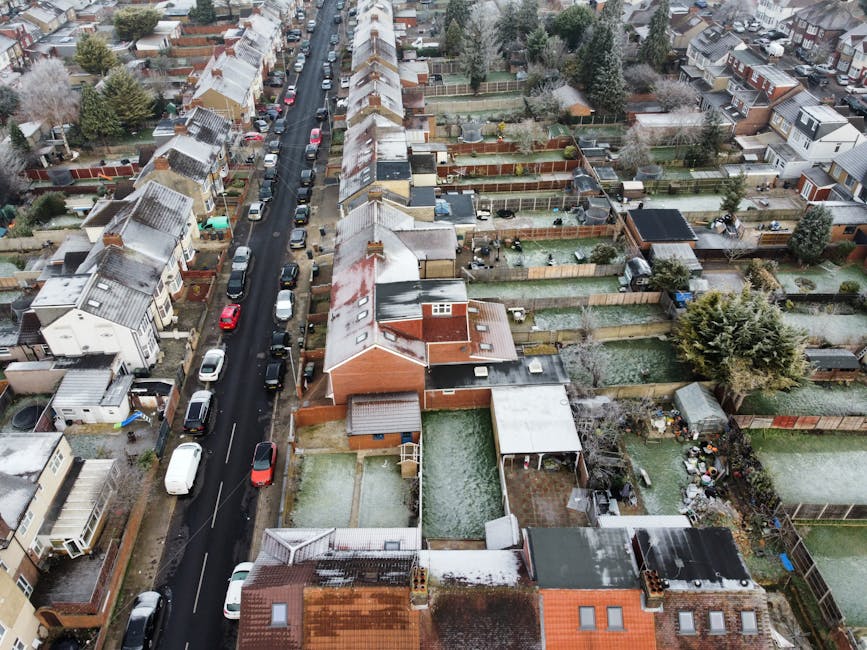 An aerial photograph of a residential neighbourhood in Haringey, showing rows of terraced houses with brick facades and tiled roofs. The street is lined with parked cars on both sides, and there are visible driveways, small gardens, and trees interspersed among the houses. The area includes green spaces and sports fields in the background, with the overall scene captured during daylight on a clear day. This view highlights typical suburban housing layout and the accessibility challenges faced during home relocation and furniture transport, as managed by Haringey Movers' removals services.