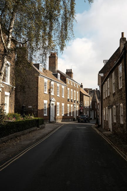 A narrow residential street in Crouch End N8 lined with brick terraced houses featuring white-framed windows and chimneys on the rooftops, with the street paved with tarmac and double yellow lines along the kerb. On the left side, a large tree with sparse green foliage extends over the street, casting shadows on the pavement. A black van is parked near the end of the street, and a small car is parked further down to the right. The scene is during the day with partly cloudy skies. This setting reflects a typical urban environment where house removals and furniture transport services, such as those provided by Haringey Movers, may be needed for home relocation or moving logistics through tight, narrow streets.