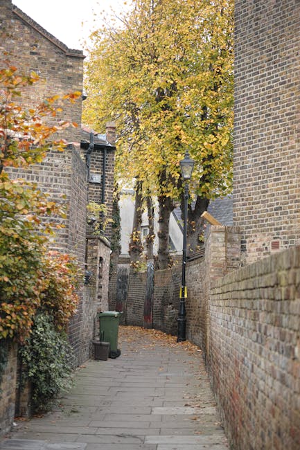 A narrow residential alleyway lined with brick walls on both sides, featuring a paved stone pathway. There is a green wheelie bin placed to the left side of the alley, near a small bush with orange leaves. An old-fashioned street lamp with a black pole and a glass lantern is situated along the right side, attached to a brick wall. Toward the end of the alley, a large deciduous tree with yellow and orange leaves extends its branches above, partially obscuring the view down the walk. The scene is illuminated by natural daylight, with some fallen leaves scattered on the pavement. This setting illustrates a typical historic street in Crouch End, N8, which can be relevant for house removals or moving services navigating narrow streets, such as those detailed in Haringey Movers' house removals tips.