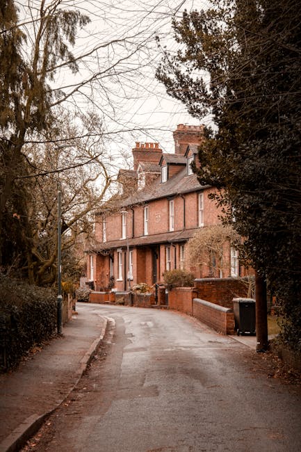A narrow residential street in Crouch End N8 lined with brick terraced houses featuring white-framed windows and chimneys on the rooftops, with the street paved with tarmac and double yellow lines along the kerb. On the left side, a large tree with sparse green foliage extends over the street, casting shadows on the pavement. A black van is parked near the end of the street, and a small car is parked further down to the right. The scene is during the day with partly cloudy skies. This setting reflects a typical urban environment where house removals and furniture transport services, such as those provided by Haringey Movers, may be needed for home relocation or moving logistics through tight, narrow streets.