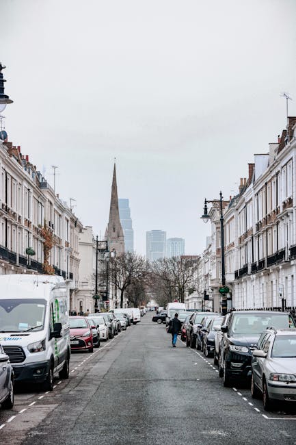 A residential street in Haringey with rows of white terraced houses featuring black iron balconies and chimney stacks, parked cars lining both sides of the narrow asphalt road, and a few pedestrians walking along the pavement. In the background, there is a tall church spire, modern high-rise buildings, and leafless trees indicating a colder season. The overcast sky creates a neutral lighting environment, suitable for depicting an urban setting typical for house removals and property relocation processes. This scene illustrates the typical environment where Haringey Movers might assist with residential furniture transport, packing, and loading during a home relocation or removal service, with a focus on logistical planning around city streets and parking arrangements.