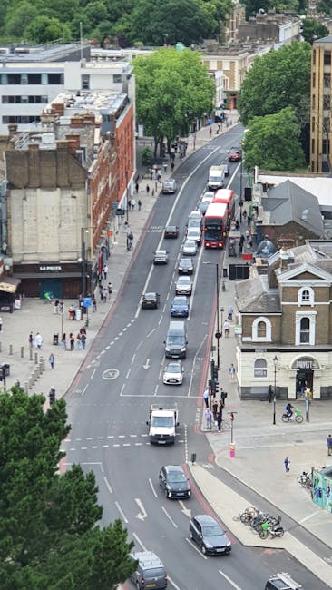An aerial view of a street in Haringey with a mix of vehicles including cars, vans, and buses traveling along a two-lane road. On one side of the street, there are pedestrians walking on the sidewalk, some of whom are waiting at crosswalks. The scene includes a variety of buildings, such as retail shops, residential houses, and apartment blocks, with lush green trees lining the street. Visible in the foreground are bicycles parked near a small roadside building, and the overall environment suggests a busy urban area suitable for house removals and furniture transport, as part of a home relocation process supported by Haringey Movers.