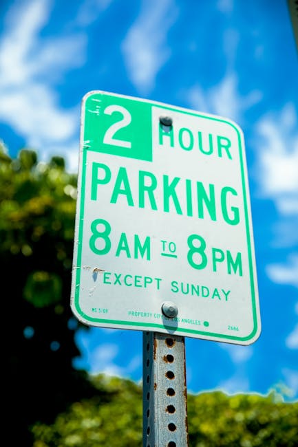 A green and white parking restriction sign mounted on a metal post outside a residential property, indicating a two-hour parking limit from 8 am to 8 pm on weekdays, with an exception for Sundays. The