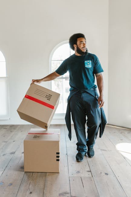 A man with an afro hairstyle and beard, dressed in a blue T-shirt and black trousers, is inside a bright, empty room with white walls and large arched windows. He is holding a cardboard box wrapped with red tape in his right hand, preparing to place or lift it onto a second identical box on the wooden floor. The scene depicts home relocation or furniture transport during a house removal, with packing materials and boxes used during the loading process. Behind him, there are no additional objects or furniture visible, emphasizing the use of packing boxes typically associated with professional move services. The natural light coming through the windows highlights the spacious interior and the focus on careful handling of packed items, which aligns with the services offered by Haringey Movers for urgent same-day removals and efficient moving logistics.