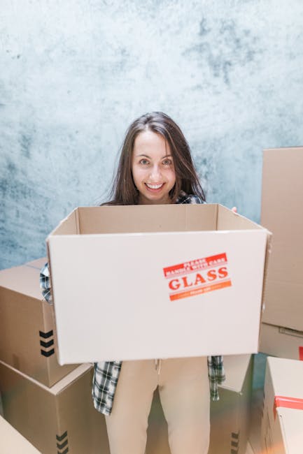A young woman with long brown hair and a cheerful expression is standing indoors in front of a plain, textured light blue wall. She is holding a large, open cardboard box with a red and white 'GLASS' warning label on one side, indicating it is intended for fragile items. Around her are several other cardboard boxes of various sizes, some sealed with packing tape and others ready for packing or transportation. The boxes are placed on the floor, and some are stacked, suggesting preparations for house removals or a home relocation process, as part of a professional moving or packing service. The environment is well-lit, emphasizing the neat and organized setup typical of packing and moving activities. This scene reflects the process of household packing and furniture transport managed by Haringey Movers, aligned with timely relocation services and efficient loading procedures within home removal projects.