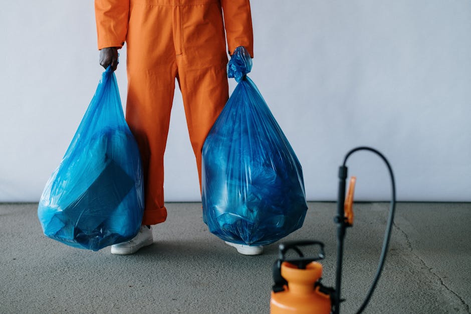 A person dressed in orange overalls and white sneakers is standing on a grey indoor floor, holding two large blue plastic garbage bags filled with household waste or items. The individual is situated