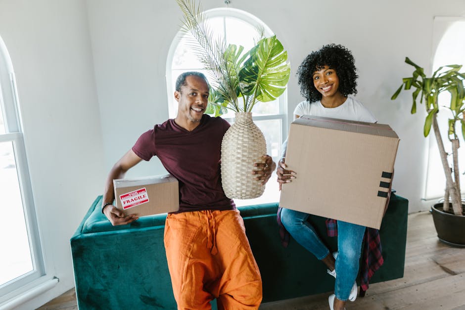 A man and a woman are inside a well-lit room with large windows, preparing for a home relocation. The man, wearing an orange pair of trousers and a maroon t-shirt, is holding a small cardboard box mar