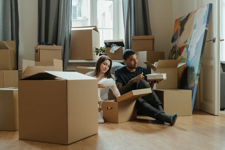 A man and a woman are inside a well-lit room with large windows, preparing for a home relocation. The man, wearing an orange pair of trousers and a maroon t-shirt, is holding a small cardboard box mar